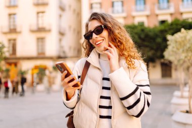Güneş gözlüklü genç bir kadının portresi. Şehir caddesinde dururken telefonla mesajlaşıyor. Neşeli bir kadın güneşli bir günde şehrin mimarisinden zevk alır. Turizm, blog yazma ve yürüme kavramı.