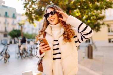 Güneş gözlüklü genç bir kadının portresi. Şehir caddesinde dururken telefonla mesajlaşıyor. Neşeli bir kadın güneşli bir günde şehrin mimarisinden zevk alır. Turizm, blog yazma ve yürüme kavramı.
