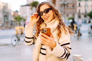 Güneş gözlüklü genç bir kadının portresi. Şehir caddesinde dururken telefonla mesajlaşıyor. Neşeli bir kadın güneşli bir günde şehrin mimarisinden zevk alır. Turizm, blog yazma ve yürüme kavramı.