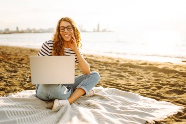 Neşeli bir kadın deniz manzarasına karşı elinde dizüstü bilgisayarla battaniyeye oturur. Genç bir kadın dizüstü bilgisayarda çalışıyor ve günbatımının tadını çıkarıyor. Bağımsızlık, doğa ve teknoloji kavramı.