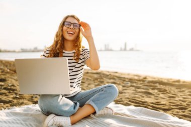 Neşeli bir kadın deniz manzarasına karşı elinde dizüstü bilgisayarla battaniyeye oturur. Genç bir kadın dizüstü bilgisayarda çalışıyor ve günbatımının tadını çıkarıyor. Bağımsızlık, doğa ve teknoloji kavramı.