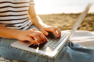 Neşeli bir kadın deniz manzarasına karşı elinde dizüstü bilgisayarla battaniyeye oturur. Genç bir kadın dizüstü bilgisayarda çalışıyor ve günbatımının tadını çıkarıyor. Bağımsızlık, doğa ve teknoloji kavramı.