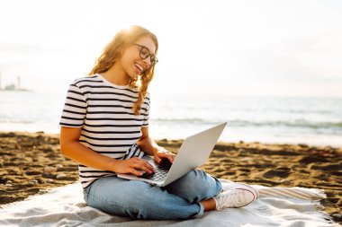 Neşeli bir kadın deniz manzarasına karşı elinde dizüstü bilgisayarla battaniyeye oturur. Genç bir kadın dizüstü bilgisayarda çalışıyor ve günbatımının tadını çıkarıyor. Bağımsızlık, doğa ve teknoloji kavramı.
