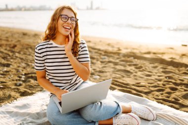 Neşeli bir kadın deniz manzarasına karşı elinde dizüstü bilgisayarla battaniyeye oturur. Genç bir kadın dizüstü bilgisayarda çalışıyor ve günbatımının tadını çıkarıyor. Bağımsızlık, doğa ve teknoloji kavramı.