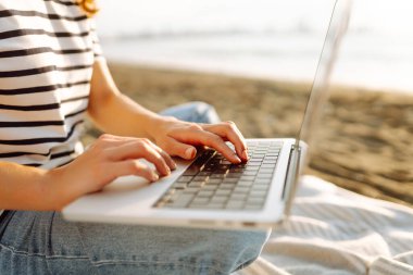 Neşeli bir kadın deniz manzarasına karşı elinde dizüstü bilgisayarla battaniyeye oturur. Genç bir kadın dizüstü bilgisayarda çalışıyor ve günbatımının tadını çıkarıyor. Bağımsızlık, doğa ve teknoloji kavramı.