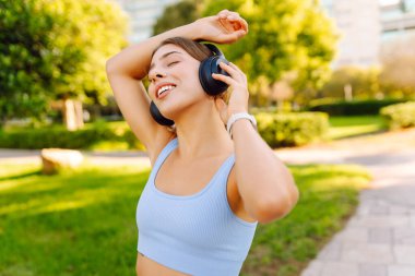 Açık mavi üniformalı mutlu bir kadın güneşli bir parkta kablosuz kulaklıkla müzik dinliyor. Kablosuz kulaklık takan atletik bir kadın açık havayı sever. Keyif ve rahatlama kavramı.