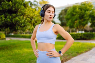 Açık mavi üniformalı mutlu bir kadın güneşli bir parkta kablosuz kulaklıkla müzik dinliyor. Kablosuz kulaklık takan atletik bir kadın açık havayı sever. Keyif ve rahatlama kavramı.