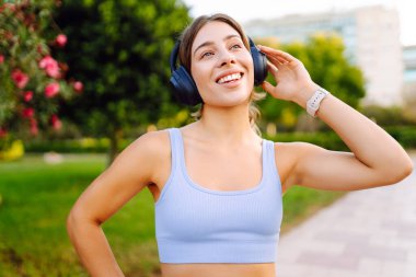 Açık mavi üniformalı mutlu bir kadın güneşli bir parkta kablosuz kulaklıkla müzik dinliyor. Kablosuz kulaklık takan atletik bir kadın açık havayı sever. Keyif ve rahatlama kavramı.