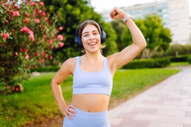 Açık mavi üniformalı mutlu bir kadın güneşli bir parkta kablosuz kulaklıkla müzik dinliyor. Kablosuz kulaklık takan atletik bir kadın açık havayı sever. Keyif ve rahatlama kavramı.