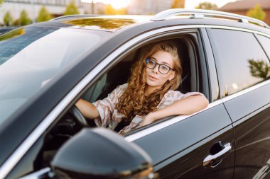 Gözlüklü, gülümseyen bir kadın arabanın direksiyonunda oturur ve pencereden dışarı bakar. Kadın sürücü güneşli günün tadını çıkarıyor. Ulaşım konsepti, taksi. Yaşam biçimi.