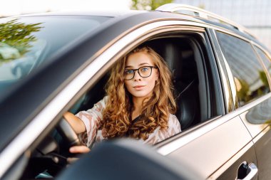 Gözlüklü, gülümseyen bir kadın arabanın direksiyonunda oturur ve pencereden dışarı bakar. Kadın sürücü güneşli günün tadını çıkarıyor. Ulaşım konsepti, taksi. Yaşam biçimi.