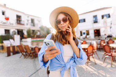 Şapkalı güzel bir kadın kalabalık bir meydanda duruyor, elinde telefon var. Güneş gözlüklü genç bir turist güneşli günün tadını çıkarıyor ve dışarıda blog yazıyor. Konsept: turizm, eğlence ve yaşam tarzı.