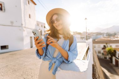 Şapkalı güzel bir kadın kalabalık bir meydanda duruyor, elinde telefon var. Güneş gözlüklü genç bir turist güneşli günün tadını çıkarıyor ve dışarıda blog yazıyor. Konsept: turizm, eğlence ve yaşam tarzı.