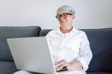 Kısa gri saçlı ve gözlüklü olgun bir kadın güneşli odada kanepede otururken dizüstü bilgisayarda çalışıyor. Güzel bir kadın rahat bir odada dizüstü bilgisayarla koltukta rahatça oturur. Mutlu yaşlanma, rahatlama kavramı.