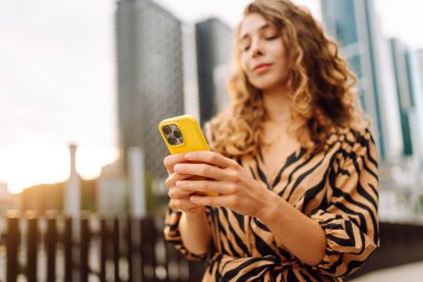 Elinde telefon olan genç bir kadının portresi metropolün arka planında duruyor. Güzel bir kadın telefonuyla mesaj atıyor ve büyük bir şehirde gezmekten zevk alıyor. Kavram: kentsel, turizm ve blog yazarlığı.