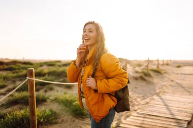 Parlak ceketli güzel bir kadın denize bakan ahşap bir patika boyunca yürüyor. Sırt çantası gün batımını sever ve dışarıda özgürce dolaşır. Yürüyüş, keyif ve doğa kavramları.