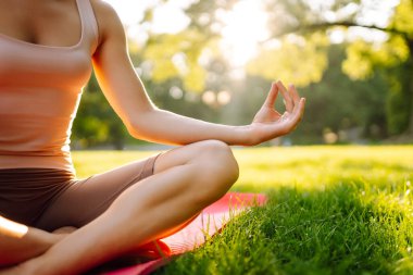Atletik giysili genç bir kadın yemyeşil parkta yoga yapıyor. Güzel bir kadın güneşli bir günün tadını çıkarır, meditasyon yapar ve paspasın üzerinde egzersiz yapar. Yoga, spor kavramları. Etkin yaşam biçimi.