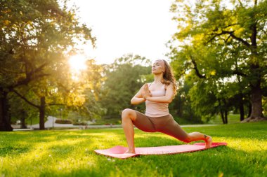 Atletik giysili genç bir kadın yemyeşil parkta yoga yapıyor. Güzel bir kadın güneşli bir günün tadını çıkarır, meditasyon yapar ve paspasın üzerinde egzersiz yapar. Yoga, spor kavramları. Etkin yaşam biçimi.