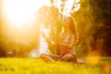 Atletik giysili genç bir kadın yemyeşil parkta yoga yapıyor. Güzel bir kadın güneşli bir günün tadını çıkarır, meditasyon yapar ve paspasın üzerinde egzersiz yapar. Yoga, spor kavramları. Etkin yaşam biçimi.
