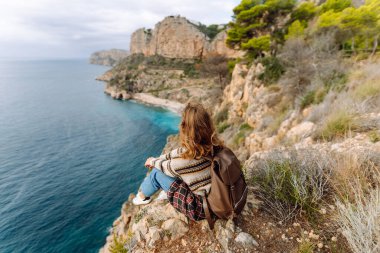 Arkadan bak. Şapka takan ve sırt çantası taşıyan kadın yürüyüşçü muhteşem deniz manzaralı bir uçurumun kenarında duruyor. Genç kadın kendini özgür hissediyor, güneşli bir günde doğanın tadını çıkarıyor. Yürüyüş kavramı, özgürlük.. 