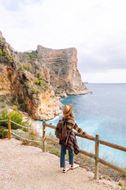 Arkadan bak. Şapka takan ve sırt çantası taşıyan kadın yürüyüşçü muhteşem deniz manzaralı bir uçurumun kenarında duruyor. Genç kadın kendini özgür hissediyor, güneşli bir günde doğanın tadını çıkarıyor. Yürüyüş kavramı, özgürlük.. 