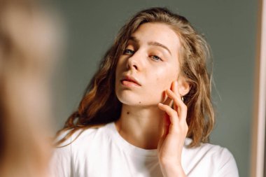 Yüzünde sivilce olan genç bir kadın güneşli bir odadaki aynaya bakıyor. Güzel kadın odada yansıyan doğal güzelliğinin tadını çıkarıyor. Tımar ve güzellik kavramı.