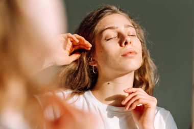 Yüzünde sivilce olan genç bir kadın güneşli bir odadaki aynaya bakıyor. Güzel kadın odada yansıyan doğal güzelliğinin tadını çıkarıyor. Tımar ve güzellik kavramı.