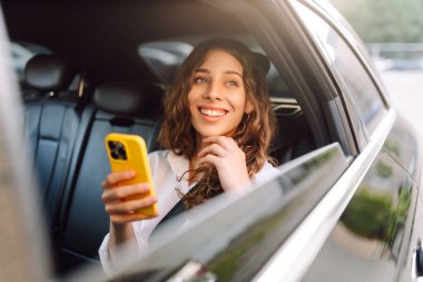 Kıvırcık saçlı güzel bir kadın güneşli bir günde elinde telefonla arabada oturuyor. Genç bir kadın arabayla gezmekten ve akıllı telefondan konuşmaktan hoşlanıyor. Kavramsal resim: ulaşım, blog yazma.