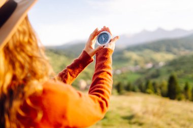 Şapkalı, elinde pusula olan genç bir kadının arka plandaki dağ manzarasına bakması. Sırt çantalı bir kadın yön bulmak için pusula kullanır ve doğada günbatımından zevk alır. Seyahat ve seyrüsefer kavramları.