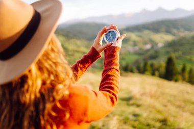 Şapkalı, elinde pusula olan genç bir kadının arka plandaki dağ manzarasına bakması. Sırt çantalı bir kadın yön bulmak için pusula kullanır ve doğada günbatımından zevk alır. Seyahat ve seyrüsefer kavramları.