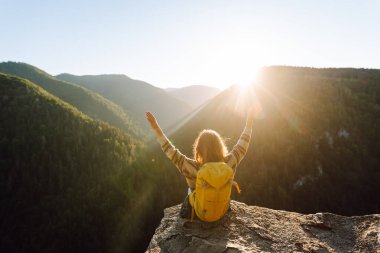 Sarı sırt çantalı genç yürüyüşçü gün batımında uçurumun kenarında duruyor. Gerçek manzara. Güzel bir kadın dağ manzarasından hoşlanır, doğanın özgürlüğünü hisseder. Özgürlük, rahatlama ve macera kavramları..