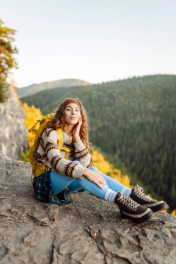 Sarı sırt çantalı genç yürüyüşçü gün batımında uçurumun kenarında duruyor. Gerçek manzara. Güzel bir kadın dağ manzarasından hoşlanır, doğanın özgürlüğünü hisseder. Özgürlük, rahatlama ve macera kavramları..