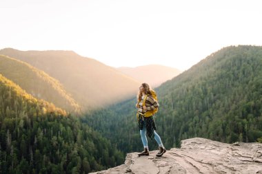 Sarı sırt çantalı genç yürüyüşçü gün batımında uçurumun kenarında duruyor. Gerçek manzara. Güzel bir kadın dağ manzarasından hoşlanır, doğanın özgürlüğünü hisseder. Özgürlük, rahatlama ve macera kavramları..