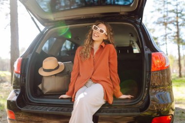 Güneş gözlüklü bir gezgin gün batımında arabanın bagajında oturur. Güzel bir kadın doğada yolculuk yapmaktan hoşlanır. Macera kavramı, ulaşım, aktif yaşam tarzı.
