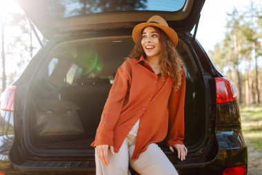 Güneş gözlüklü bir gezgin gün batımında arabanın bagajında oturur. Güzel bir kadın doğada yolculuk yapmaktan hoşlanır. Macera kavramı, ulaşım, aktif yaşam tarzı.