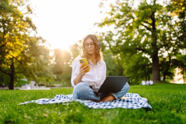 Dizüstü bilgisayarı ve telefonu olan genç bir kadın gün batımında Green Park 'ta halının üzerinde oturuyor. Serbest çalışan bir kadın, telefonu olan bir öğrenci, dışarıda bir laptopta çalışıyor ya da çalışıyor. Uzak çalışma konsepti, teknoloji.