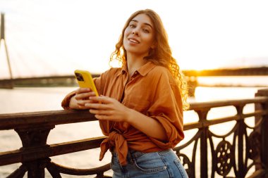 Gün batımında setin üzerinde telefonu olan genç bir kadının portresi duruyor. Güzel bir kadın telefonuyla mesaj atıyor ve dışarıda dolaşmaktan hoşlanıyor. Kavram: teknoloji, şehir, blog. Yaşam biçimi.