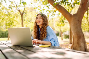 Dizüstü bilgisayarı olan neşeli bir kadın güneşli bir parktaki masada oturuyor. Dişi bir serbest çalışan güneşli bir günün tadını çıkarır ve dizüstü bilgisayarıyla çalışır. Bağımsız kavram, eğitim.