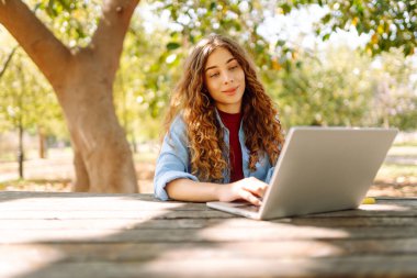 Dizüstü bilgisayarı olan neşeli bir kadın güneşli bir parktaki masada oturuyor. Dişi bir serbest çalışan güneşli bir günün tadını çıkarır ve dizüstü bilgisayarıyla çalışır. Bağımsız kavram, eğitim.