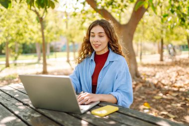 Dizüstü bilgisayarı olan neşeli bir kadın güneşli bir parktaki masada oturuyor. Dişi bir serbest çalışan güneşli bir günün tadını çıkarır ve dizüstü bilgisayarıyla çalışır. Bağımsız kavram, eğitim.