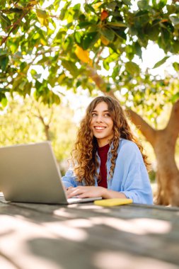 Dizüstü bilgisayarı olan neşeli bir kadın güneşli bir parktaki masada oturuyor. Dişi bir serbest çalışan güneşli bir günün tadını çıkarır ve dizüstü bilgisayarıyla çalışır. Bağımsız kavram, eğitim.