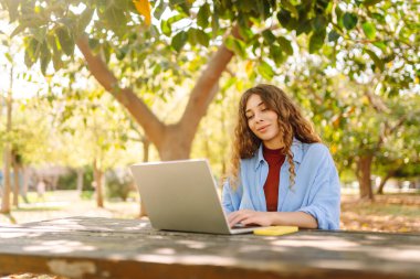 Dizüstü bilgisayarı olan neşeli bir kadın güneşli bir parktaki masada oturuyor. Dişi bir serbest çalışan güneşli bir günün tadını çıkarır ve dizüstü bilgisayarıyla çalışır. Bağımsız kavram, eğitim.