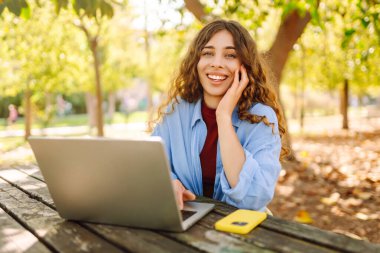 Dizüstü bilgisayarı olan neşeli bir kadın güneşli bir parktaki masada oturuyor. Dişi bir serbest çalışan güneşli bir günün tadını çıkarır ve dizüstü bilgisayarıyla çalışır. Bağımsız kavram, eğitim.