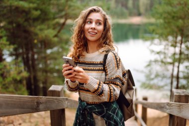 Sırt çantası ve telefonu olan bir gezginin portresi gölün kenarındaki ahşap bir yolda duruyor. Tatlı bir kadın blogu ya da açık havada telefonundaki mesajlar. Seyahat ve teknoloji kavramları.