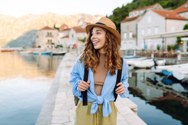 Şapkalı güzel bir turist ve şafak vakti rıhtımda sırt çantasıyla geziniyor. Genç kadın güneş ışınlarında deniz manzarasını seviyor. Seyahat, rahatlama, aktif yaşam tarzı kavramları.