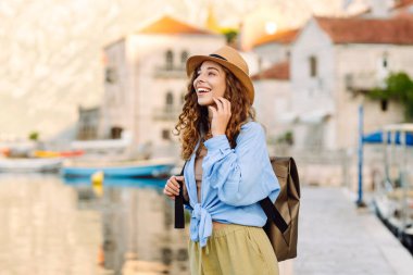 Şapkalı güzel bir turist ve şafak vakti rıhtımda sırt çantasıyla geziniyor. Genç kadın güneş ışınlarında deniz manzarasını seviyor. Seyahat, rahatlama, aktif yaşam tarzı kavramları.