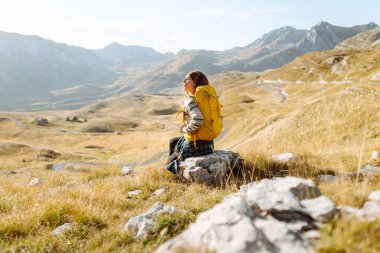 Sarı sırt çantalı güzel bir bayan yürüyüşçü dağ sırtında durup günbatımını izliyor. Genç kadın yolculuğun tadını çıkarıyor ve gün batımının özgürlüğünü hissediyor. Yürüyüş, özgürlük ve aktif yaşam tarzı kavramı.