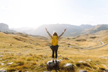 Sarı sırt çantalı güzel bir bayan yürüyüşçü dağ sırtında durup günbatımını izliyor. Genç kadın yolculuğun tadını çıkarıyor ve gün batımının özgürlüğünü hissediyor. Yürüyüş, özgürlük ve aktif yaşam tarzı kavramı.