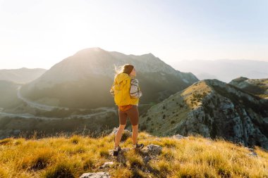 Sarı sırt çantalı güzel bir bayan yürüyüşçü dağ sırtında durup günbatımını izliyor. Genç kadın yolculuğun tadını çıkarıyor ve gün batımının özgürlüğünü hissediyor. Yürüyüş, özgürlük ve aktif yaşam tarzı kavramı.