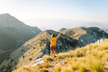 Sarı sırt çantalı güzel bir bayan yürüyüşçü dağ sırtında durup günbatımını izliyor. Genç kadın yolculuğun tadını çıkarıyor ve gün batımının özgürlüğünü hissediyor. Yürüyüş, özgürlük ve aktif yaşam tarzı kavramı.